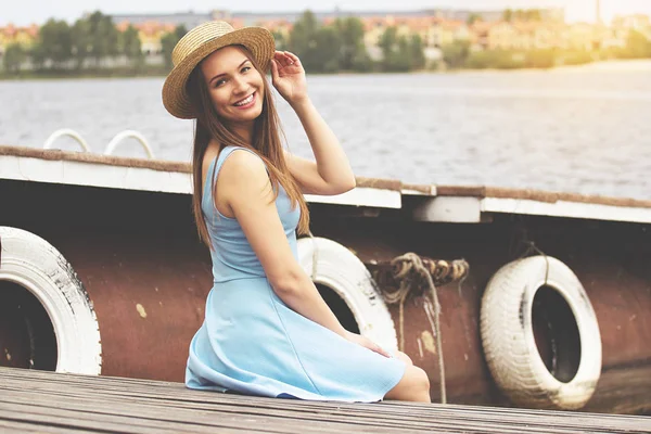 Shining beauty. Side view horizontal shot of attractive young woman in dress adjusting hat on her head and smiling while sitting on wooden pier.