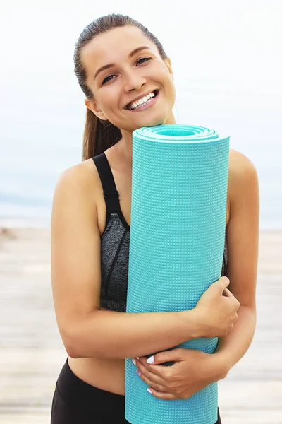 Training für gute Laune. Porträt einer schönen und jungen Frau in Sportkleidung, die in die Kamera blickt und lächelt, während sie Yogamatte am Strand umarmt. — Stockfoto