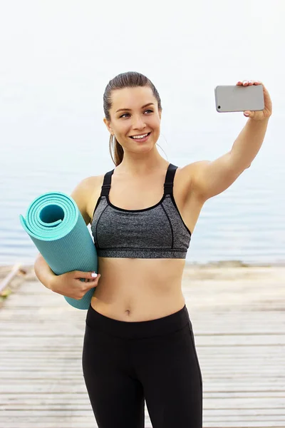 Sport-Selfie. schöne junge Fitness-Frau hält ihr Handy in der Hand und lächelt, während sie ein Selfie mit Yogamatte auf einem Holzsteg am Meer macht. — Stockfoto