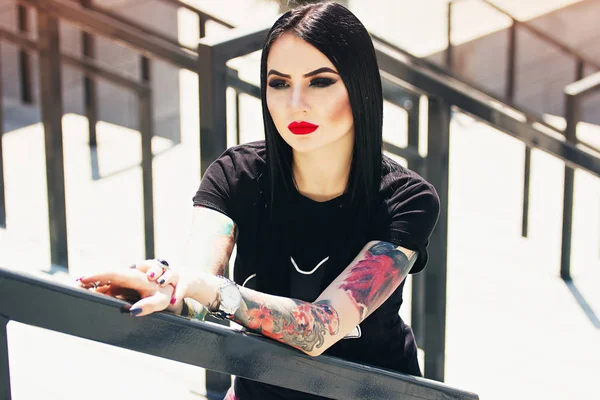 Moda de tatuagem. Retrato de menina hipster tatuado na moda com lábios vermelhos posando contra fundo urbano . — Fotografia de Stock