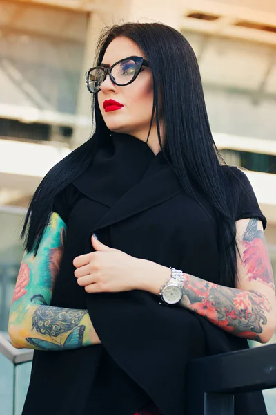 Gothic beauty. Portrait of attractive tattooed hipster girl with red lips posing to camera while standing against urban background.