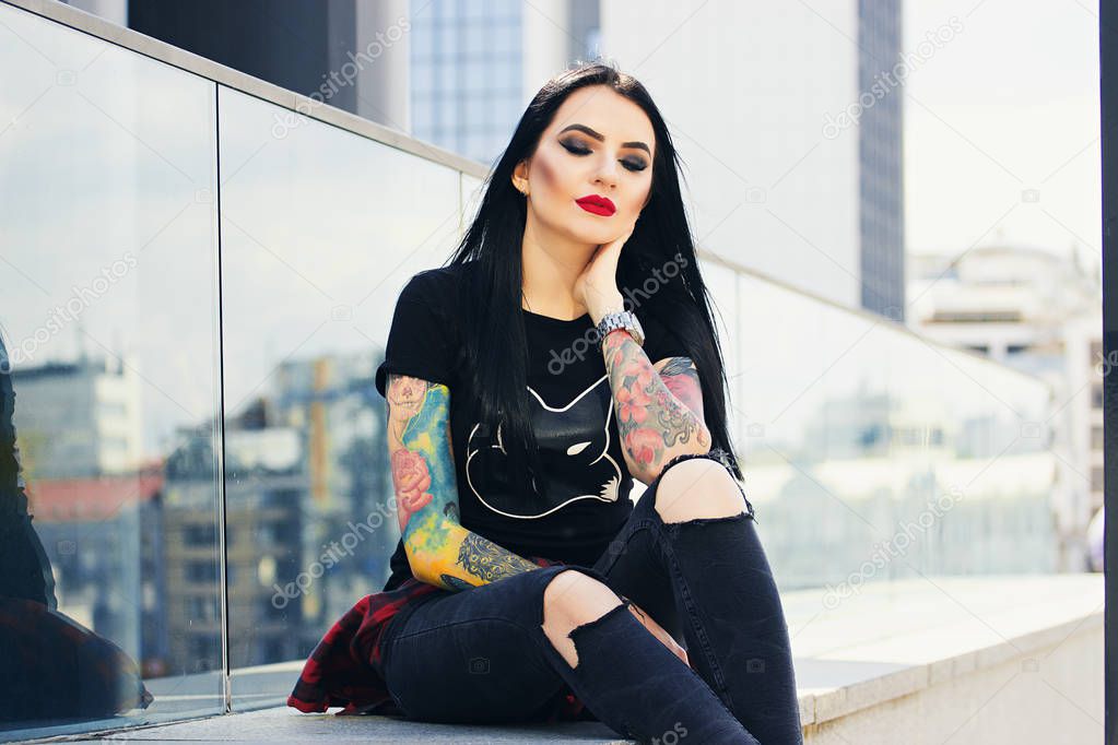 Gothic beauty. Portrait of attractive tattooed hipster girl with red lips posing to camera while sitting against urban background.