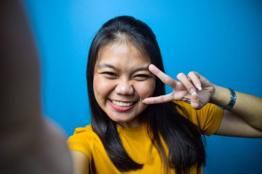 Mavi izole edilmiş geçmişi olan güzel Asyalı kadınların portresi, selfie çeken ve barış ya da zafer işareti yapan gülümsemeler.