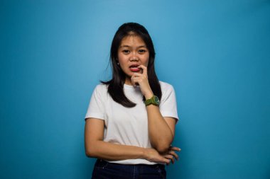 Mavi izole edilmiş arka planı olan beyaz tişört giyen güzel Asyalı kadınların portresi kameraya bak ve tırnak ye, gergin bir jest yap.