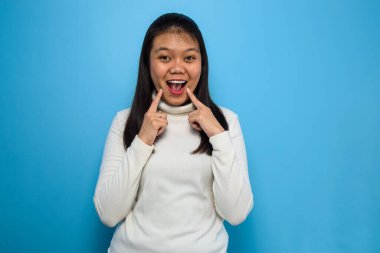 Genç Asyalı güzel kadınların portresi mavi izole edilmiş arka planı olan beyaz tişörtler kullanıyor, parmakları neşeli gülümsemeyi işaret ediyor ve zorluyor.