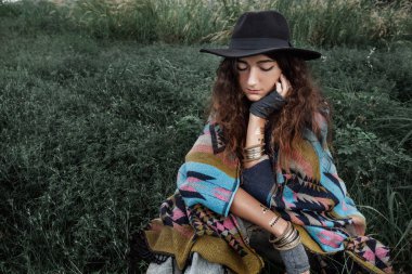 Yeşil alanında oturup panço ve şapka giyen güzel boho kadın