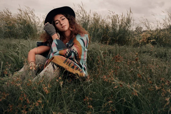 Yeşil alanında oturup panço ve şapka giyen güzel boho kadın — Stok fotoğraf