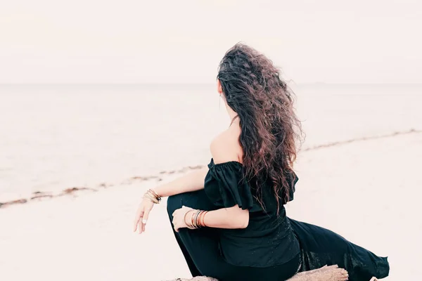 Jovem elegante em vestido preto sentado na praia ao sol — Fotografia de Stock