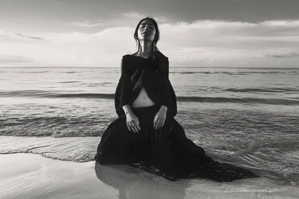 Modèle jeune femme à la mode assis sur le sable dans l'eau au coucher du soleil — Photo