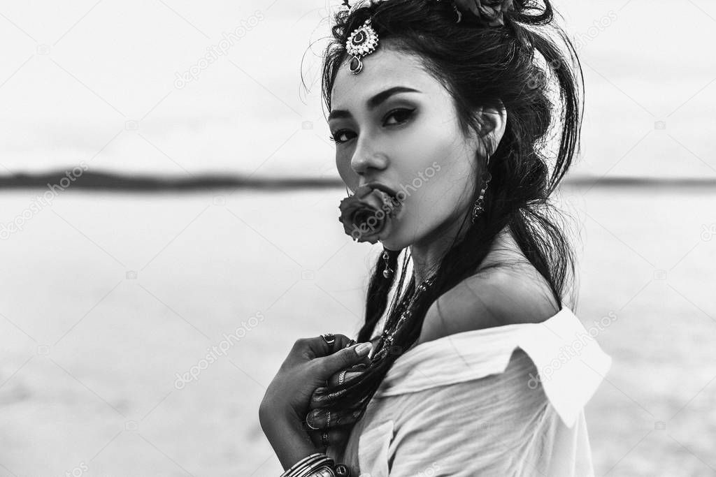 oriental stylish boho girl holding flower in mouth on the beach at sunset. black and white portrait