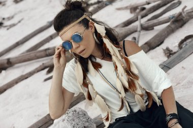 Güneş gözlüklü genç bir kadın ve kumsalda sırt çantalı beyaz bir gömlek.