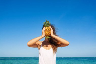 Sahilde ananasla poz veren zarif genç bir kadın.