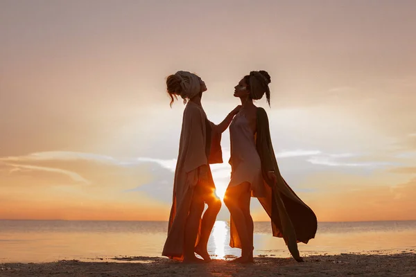 สาวสวยสองคนในผ้าพันคอบนชายหาดตอนพระอาทิตย์ตก — ภาพถ่ายสต็อก