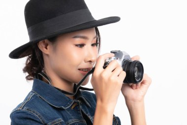 Siyah şapkalı ve kot ceketli güzel bir bayan turist, beyaz arka planda izole edilmiş stüdyo çekimlerinde mutluluk içinde klasik bir kamerayla fotoğraf çekmek için ayakta duruyor..