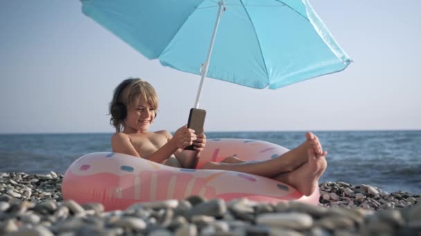 Χαριτωμένο παιδί ακούγοντας μουσική στο έξυπνο τηλέφωνο στην παραλία — Αρχείο Βίντεο
