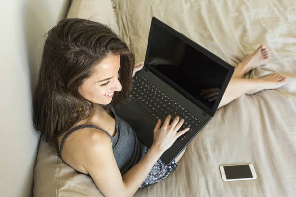 Wanita muda yang bahagia duduk di tempat tidur menggunakan laptop — Stok Foto