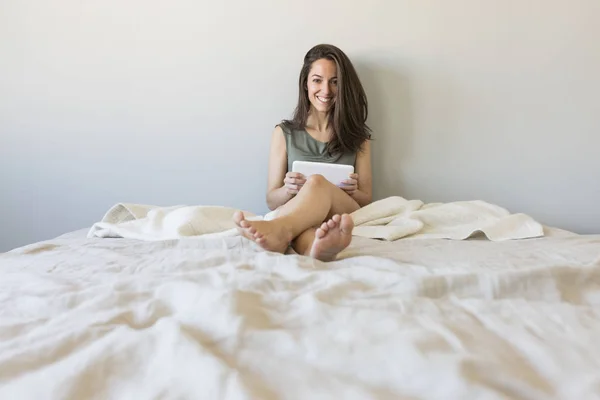 Wanita muda yang cantik tersenyum dan menggunakan tabletnya di tempat tidur — Stok Foto