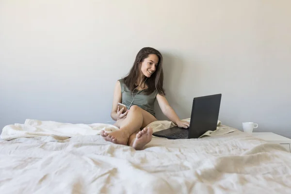 Szczęśliwy piękna młoda kobieta siedzi na łóżku za pomocą laptopa — Zdjęcie stockowe