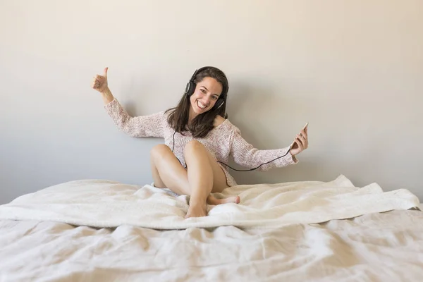 Wanita muda yang bahagia mendengarkan musik di tempat tidur dengan ponsel — Stok Foto