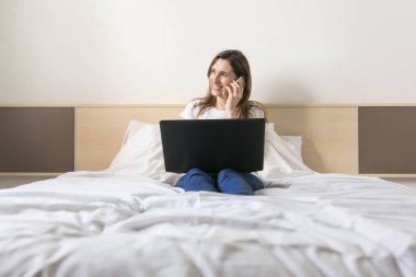 laptop ve konuşma kullanarak yatağın üstüne oturan mutlu güzel genç kadın