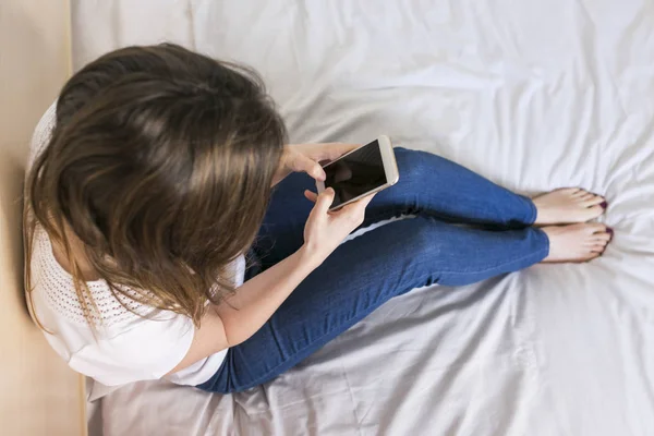 Wanita muda duduk di tempat tidur dan menggunakan ponsel di rumah. Dayti — Stok Foto