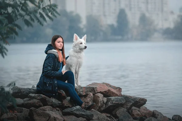 Śliczna Nastolatka Dziewczyna z jej psa rasy mieszanej, siedząc na kamieni na brzegu rzeki, z widokiem na miasto na jesieni — Zdjęcie stockowe