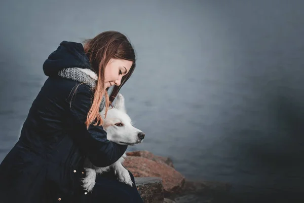 Beautiful teenager girl hugging her white dog on the bank of the river autumn morning — Stock Photo, Image