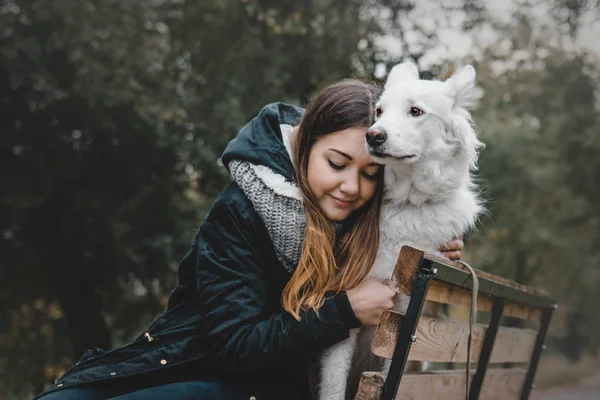 Beautiful teenager girl hugging her white dog on the bank of the river autumn morning — Stock Photo, Image