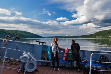 Listvyanka, Russia - June 26, 2019: People look at the village Port Baikal from a ferry, Russia. Horizontal day view of the high shore, green forest, clear lake water