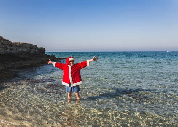 Felice uomo anziano in costume Babbo Natale è in piedi sulla spiaggia — Foto Stock