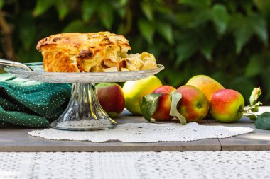 Cam tezgahta ev yapımı elmalı turta. Yeşil yaprakların doğal arka planında beyaz, kaliteli bir peçete var. Güneşli bir bahçe ve organik ürünlerle sağlıklı bir yaşam tarzı..