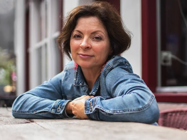 Modern woman of 50-60 years old, in casual clothes, sitting at a table in a cafe on a blurred background.