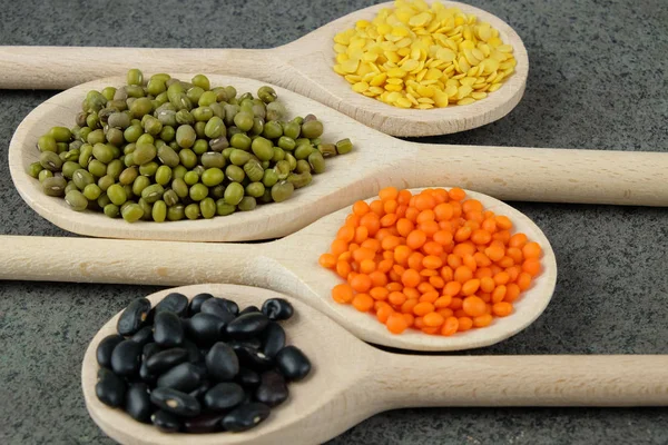 Vários tipos de lentilhas e feijão preto em colheres de madeira . — Fotografia de Stock