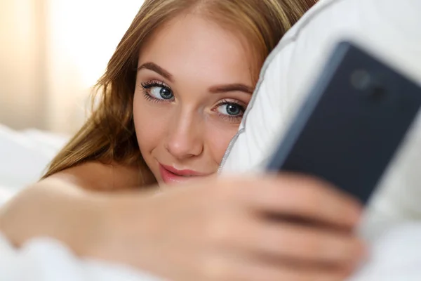 Jonge mooie blonde vrouw liggend in bed holding cellphone — Stockfoto