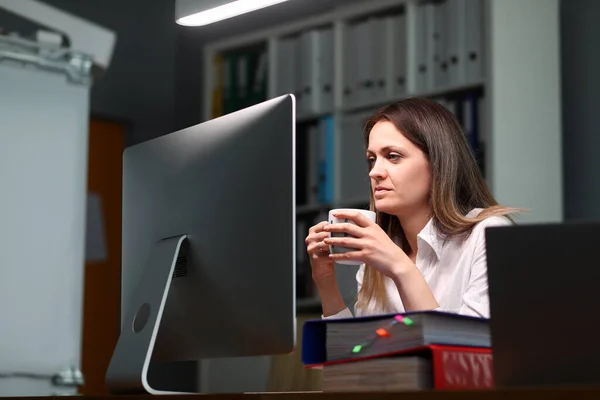 Frau hält Becher in der Hand und blickt intensiv auf Monitor — Stockfoto