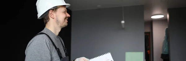Jovem reparador sorridente em pé em apartamento vazio e segurando o plano de construção nas mãos — Fotografia de Stock
