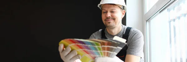 Young repairman in helmet sitting in flat and choosing color for interior — Stockfoto