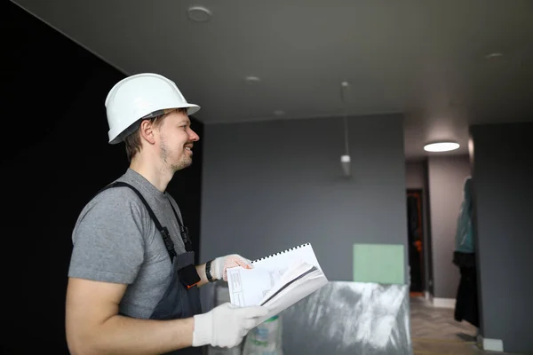 Jovem reparador sorridente em pé em apartamento vazio e segurando o plano de construção nas mãos — Fotografia de Stock