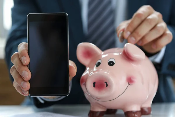 In hands smartphone, man puts coin in piggy bank — Stock Photo, Image