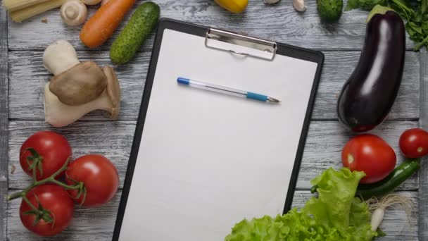 Rollo de la pluma en el portapapeles en blanco en la mesa de cocina con verduras — Vídeos de Stock