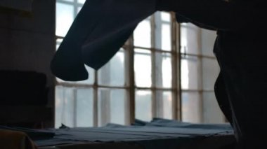 Kadın kesilmemiş bir parça süet kumaşı alıp sallıyor ve masanın üzerine seriyor.