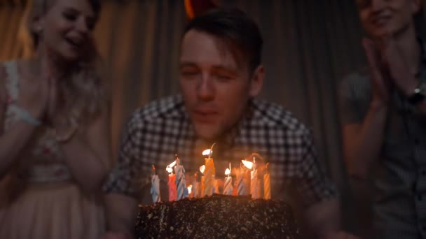 Young man blows out candles on a festive cake with friends near. — Stock Video
