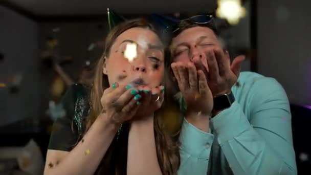 Młoda para zabawia się na imprezie, nosi imprezowe kapelusze, dmucha konfetti — Wideo stockowe