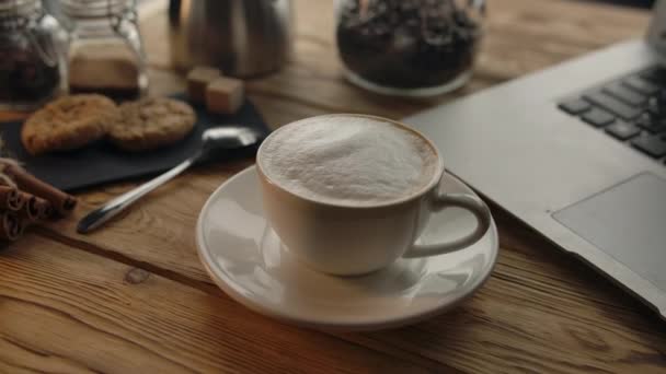 Una taza de café en una mesa de madera junto a un portátil — Vídeo de stock