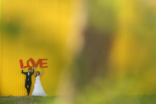 Alegre pareja nupcial cerca de la pared amarilla sosteniendo letras de AMOR — Foto de Stock