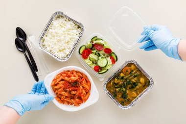 Tek kullanımlık konteynırlarda servis edilen yemekler: pirinç, sebze körisi, domates soslu İtalyan makarnası ve taze sağlıklı salata. Ve tıbbi eldivenli eller. Karantina
