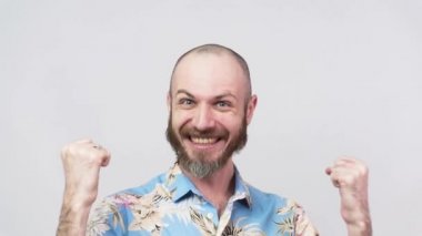 Hawaii gömleği giyen mutlu bir adam beyaz arka plandaki zaferi yavaş çekimde kutluyor. Başarı, zafer, zafer, kazanma ve kutlama duyguları.