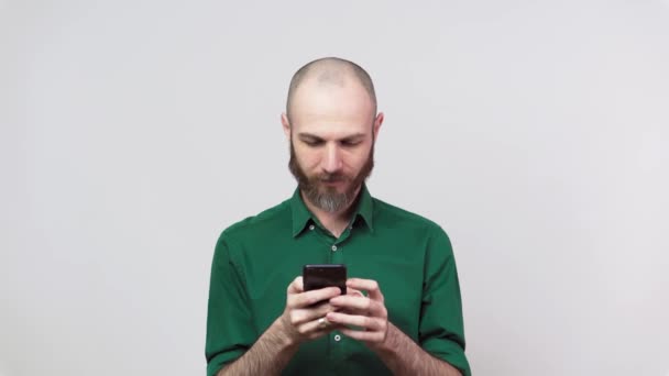 Knappe gelukkige man met een baard die een mobiele telefoon over een witte achtergrond gebruikt. Mensen oprechte emoties, levensstijl concept. Kijk verrast met behulp van mobiele telefoon. — Stockvideo