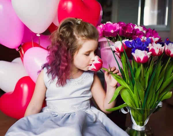 Pequena menina bonita com cabelo encaracolado rosa em um vestido de prata em tulipas vermelhas e com bolas multi-coloridas — Fotografia de Stock