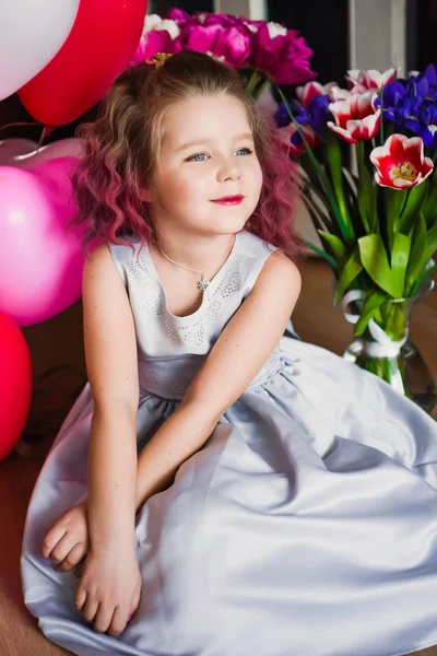 Pequena menina bonita com cabelo encaracolado rosa em um vestido de prata em tulipas vermelhas e com bolas multi-coloridas — Fotografia de Stock